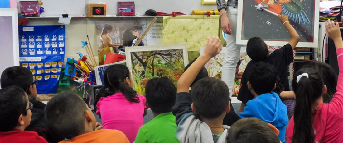 A group of young students sit on the floor in a classroom, some raising their hands, as a teacher holds up illustrated artwork.