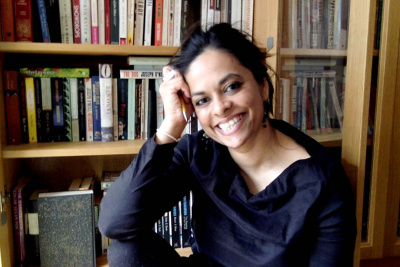 Color photograph of architect Sofia Karim smiling into the camera, head in her hands while she holds a pencil, and sitting in front of a full bookcase. 