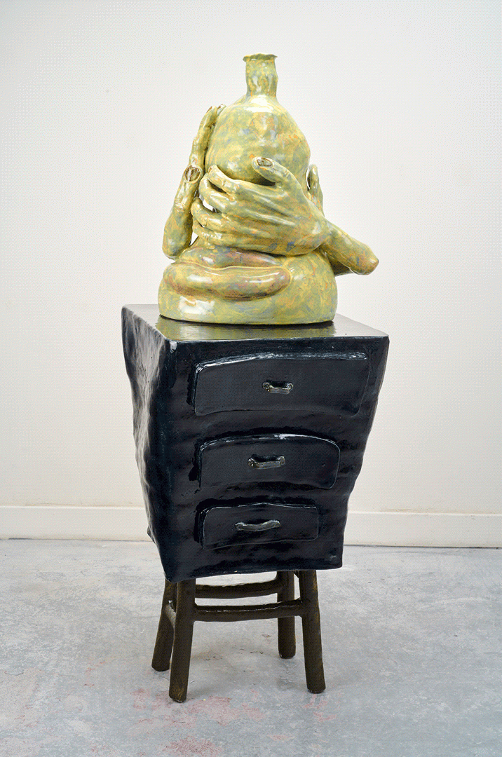 A mint green and yellow vessel rests on a stand with 3 sealed drawers. The stand's blue-green texture is lumpy. It is top heavy, getting narrower as it is closer to the floor. The vessel resembles a short stout abstract face, as a hand curls from the right to cover hidden eyes. 