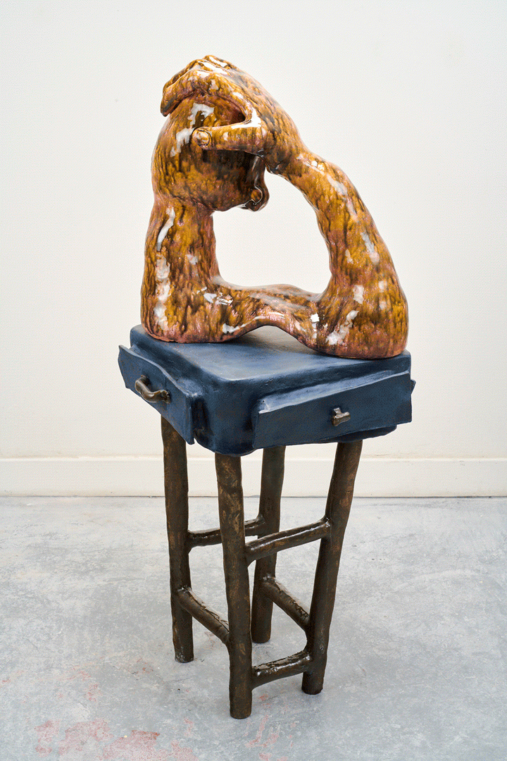 A caramel orange glazed arm reaches up and rests atop the side profile of a head, creating a circle. This form sits atop a blue wobbly stand with brown thin legs—the drawers of the stand peaking open.