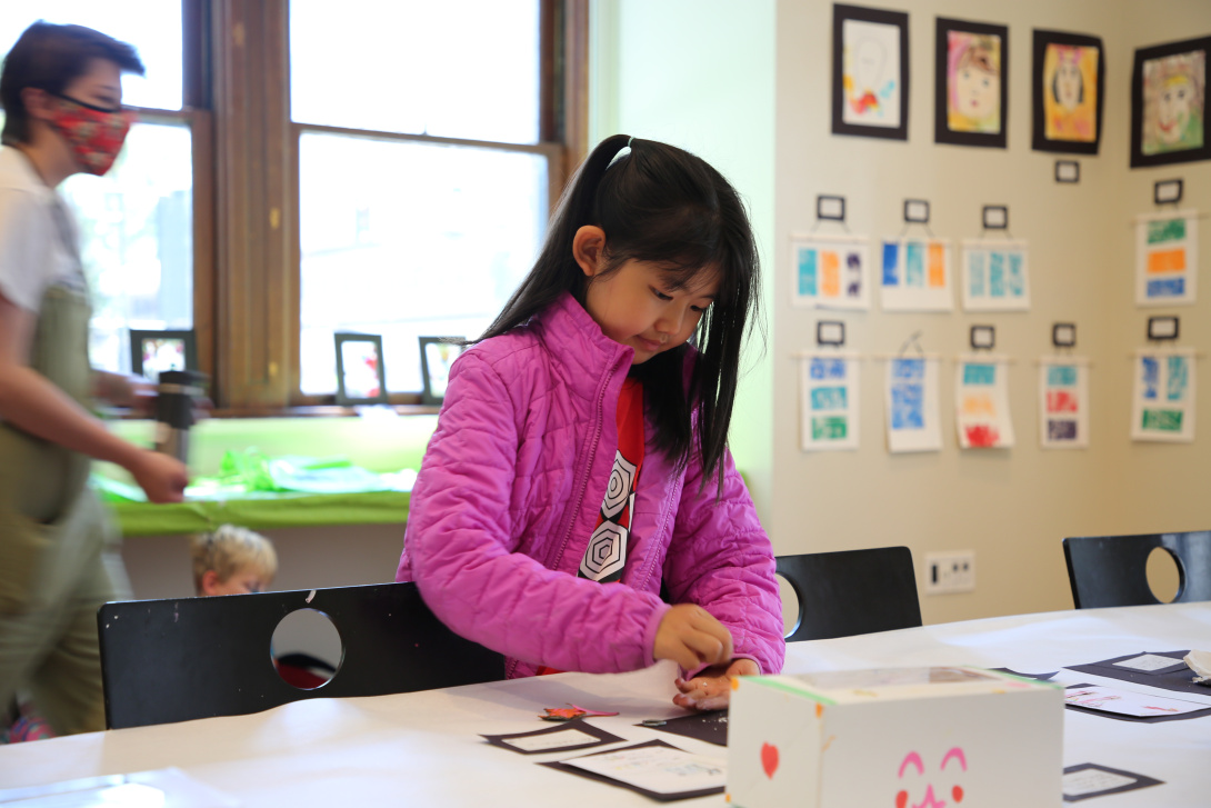 In a room filled with children’s art, a young Asian girl places something on a table. In the background is an adult wearing a smock.