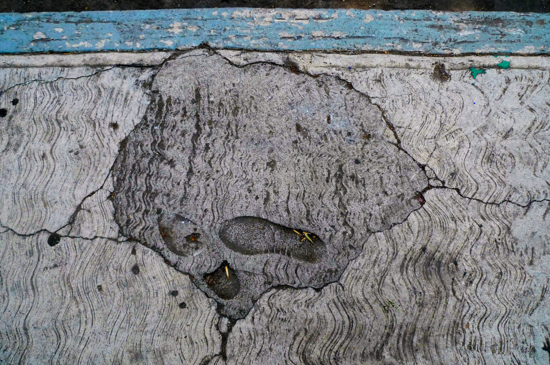 A view from above of footstep imprints left in a cement sidewalk. The cement is cracked and various shades of dark gray. At the top is a blue line that signals the edge of the sidewalk and indicates that the space next to it is reserved for disabled parking.