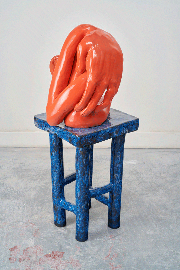 A fluorescent orange vessel rests on a tall, blue marble-glazed stool that appears wobbly with legs that get thinner as they reach the floor. The vessel resembles an abstracted arm curling in on itself, the overly large hand hiding most of itself. 