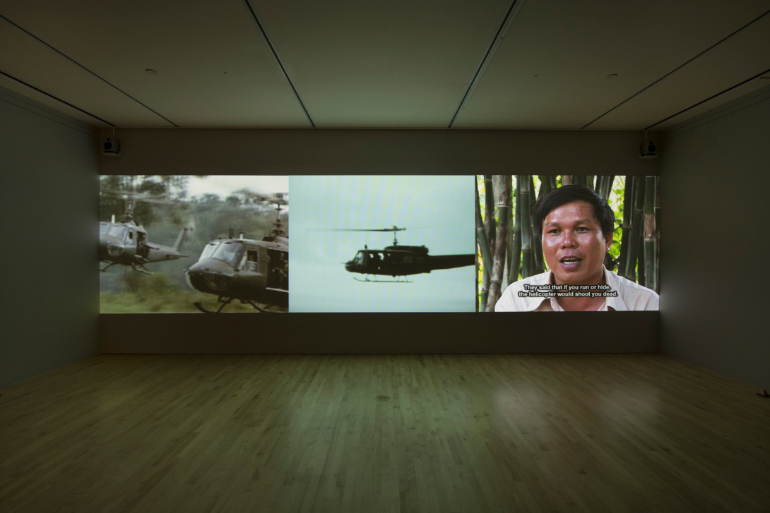 A video installation divided into three sections. A flying military helicopter is in the center, two military helicopters flying low in a forest is on the left, while a man with overlaid text, “They said that if you run or hide, the helicopter would shoot you dead” is on the right.