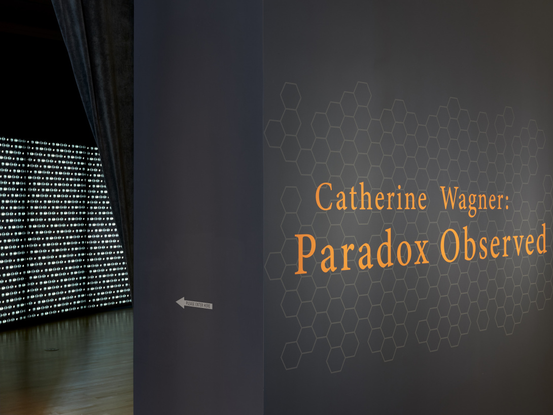 Pumpkin orange text on a graphite colored wall reads, "Catherine Wagner: Paradox Observed." The exhibition text sits on a hexagonal pattern. To the left, an arrow points to a dark installation space where a large wall of lights awaits.