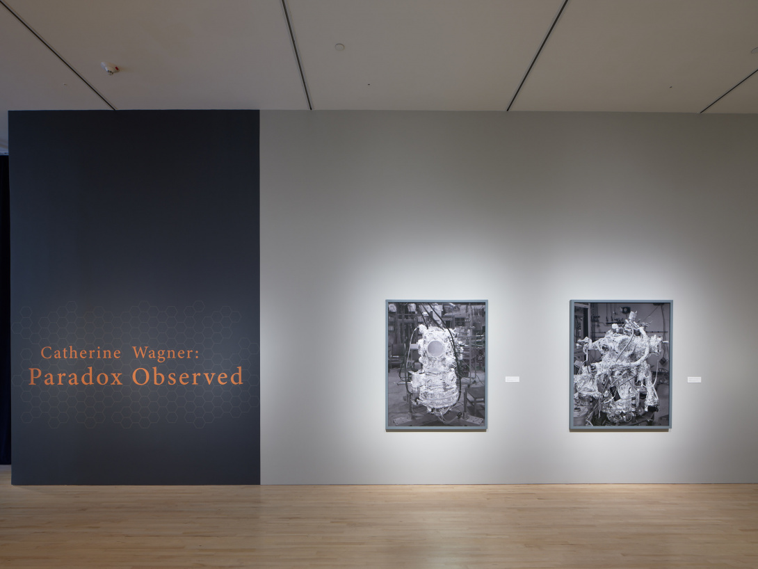 Pumpkin orange text on a graphite colored wall reads, "Catherine Wagner: Paradox Observed." Two rectangular grey scale images hang side by side. The images show foil-wrapped scientific machines, both connected to tubing and hoses.