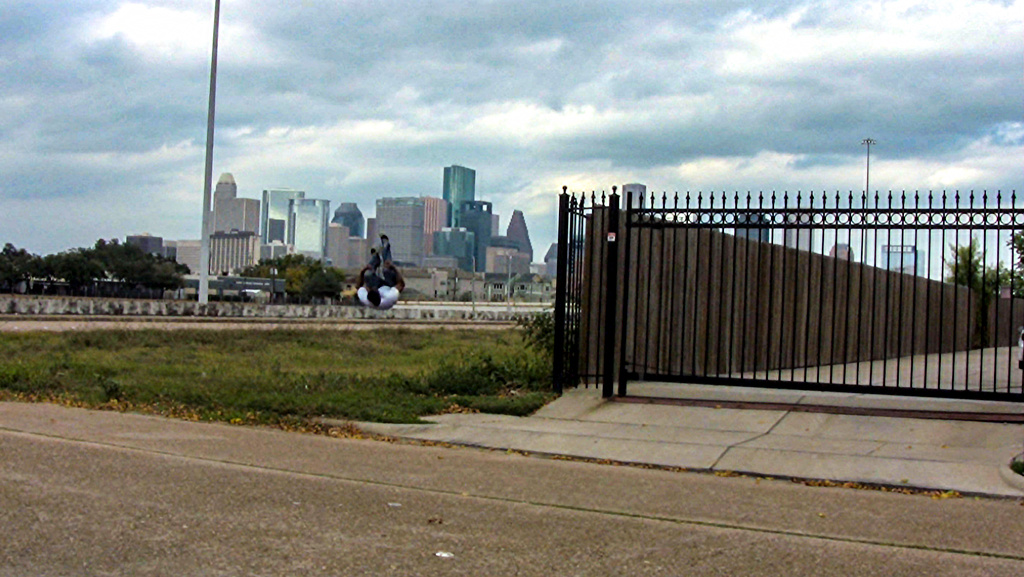 In front a cityscape, a black person is mid-air, as though he is doing a back-flip or falling from the sky. A large metal fence is to the left, with green grass below the jumper, and in front of him is a cement street and block. Blue skies look ominous,