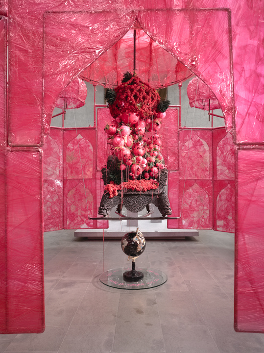 A close-up of a pink fragile (perhaps paper) sculpture in which we peer through the pink doorway at an ancient throne laden with what appears to be pink pomegranates or some other fruit. The entire display floats off the ground above a marble globe. 