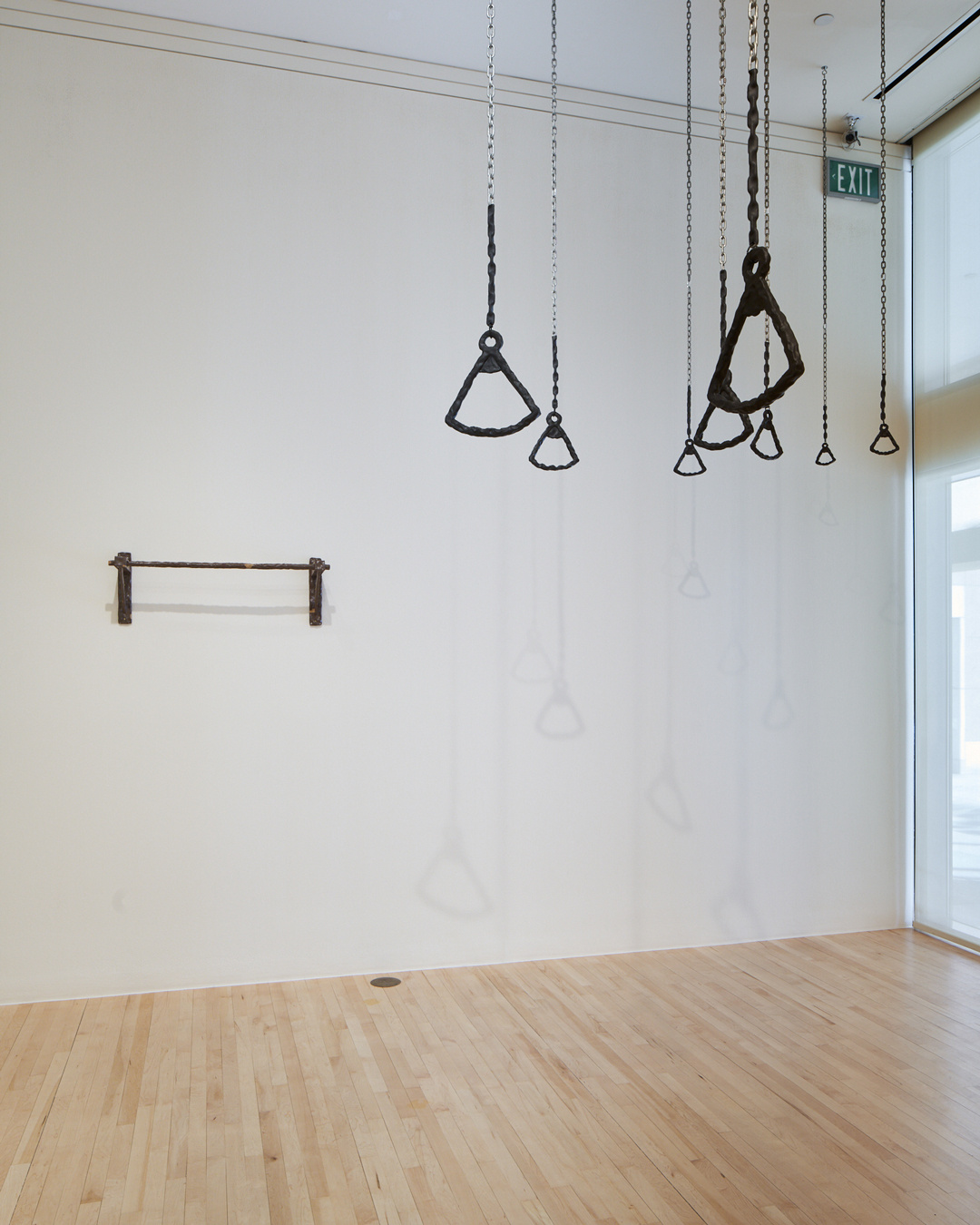 An off-white gallery wall is barren except for a small, metal pull-up bar positioned on the wall's left, midway to the ceiling. In the upper right quadrant, eight black trapeze pull up bars hang from chains on the ceiling and cast faint shadows onto the gallery wall.