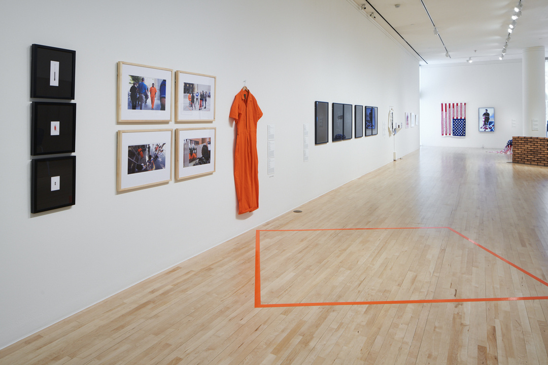 A gallery with an orange prison jumpsuit hangs on the wall, with 3 black framed images to the left of it along with four color framed prints. In the background is an American flag and a partial brick wall with other framed works that are indistinguishable.