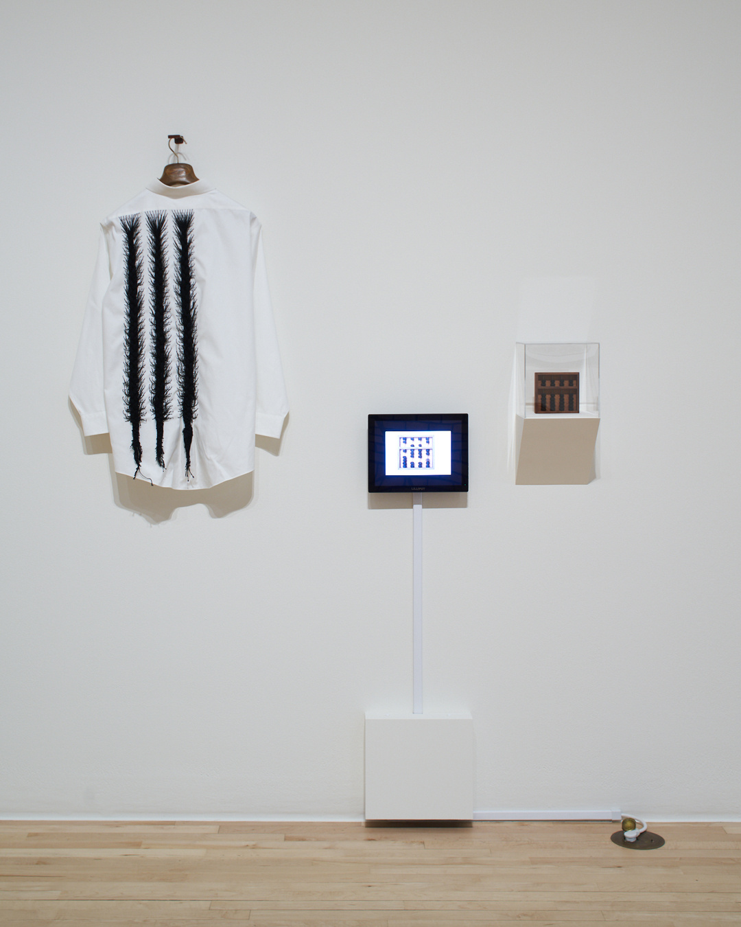 An off-white wall displays a long-sleeved white shirt, a tablet screen, and a small artwork encased in a plexiglass cube. The shirt has three black, feathered lines on its left side that extends vertically from the shoulders down to the bottom.