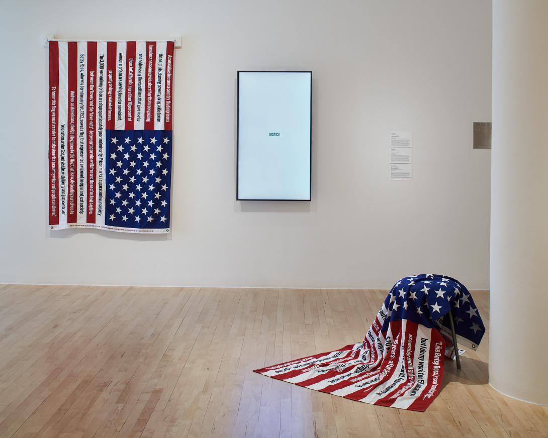 Two American flags embroidered with text. The left flag is pinned to the wall. The right flag is gently draped across a stool, as though someone just stopped embroidering. Between the 2 flags is a large TV with a white screen and small text reading "Justice."