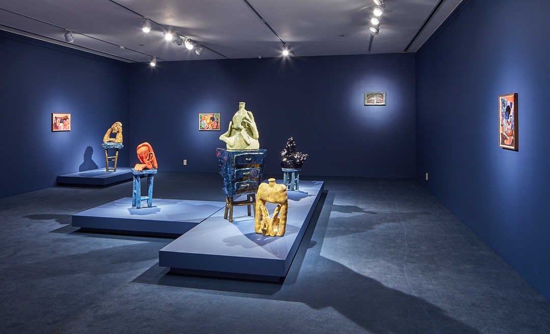 A gallery shot with dark blue walls, dark blue plinths, and dark blue carpeting. Spotlights highlight several small paintings. The raisers in the center of the room feature several anthropomorphized sculptures that are somewhere between vase and model.