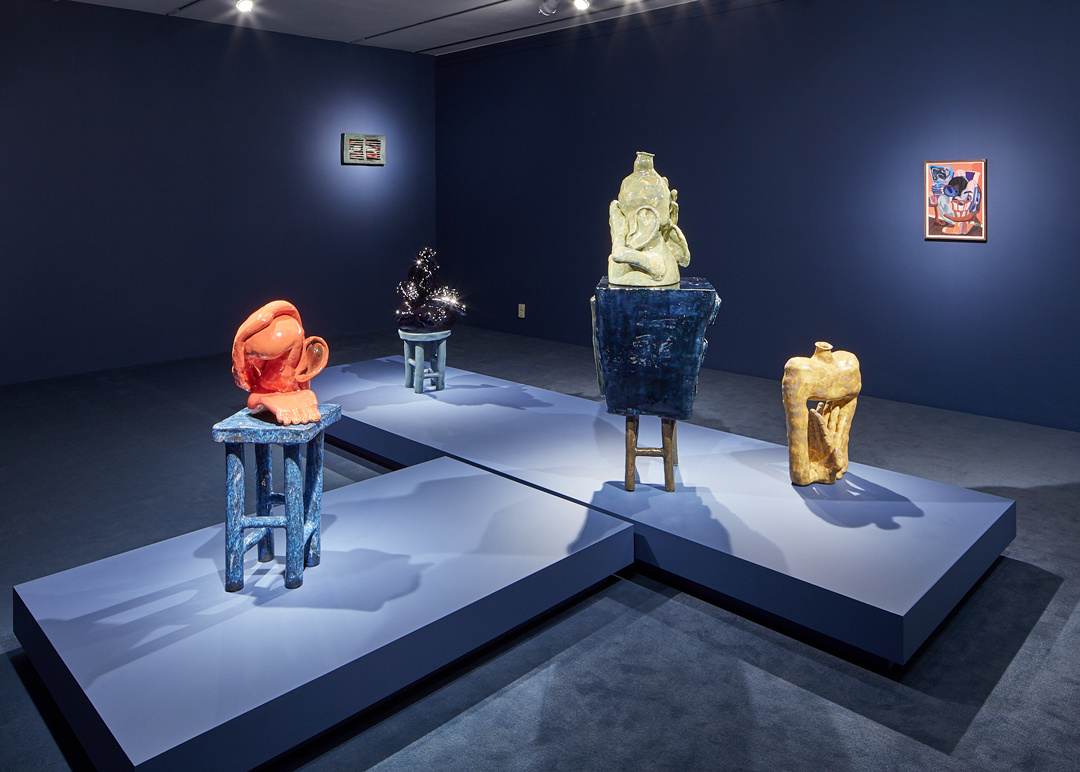 Dark blue painted gallery with a T-shaped pedestal featuring three sculptures on stools and one standing directly on the pedestal.  The sculptures have spotlights illuminating them. Two spotlighted paintings are on the walls.