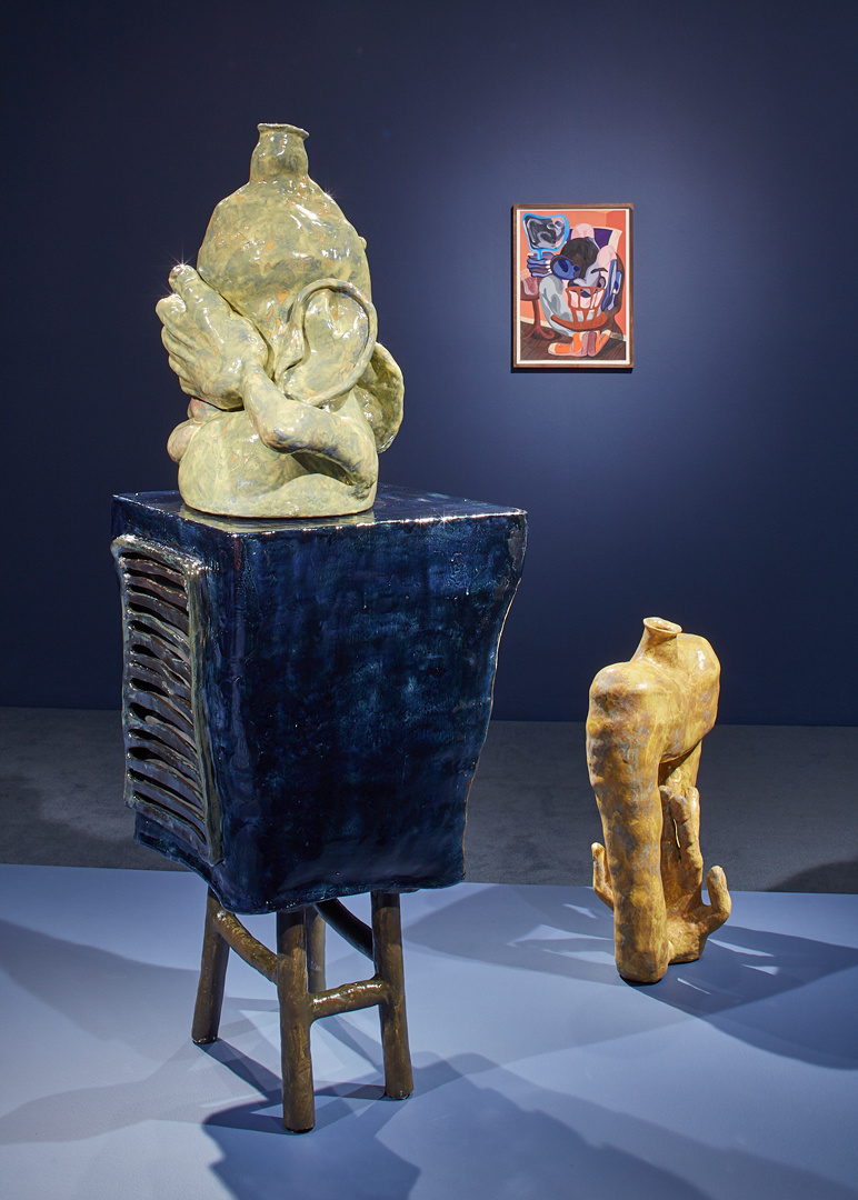 Multiple sculptures are in a blue gallery. The sculptures have a melting quality, like they are breathing and moving. One ceramic in  has a large ear and hand covering what could be its eyes. A bold painting of warm colors hangs in the background. 