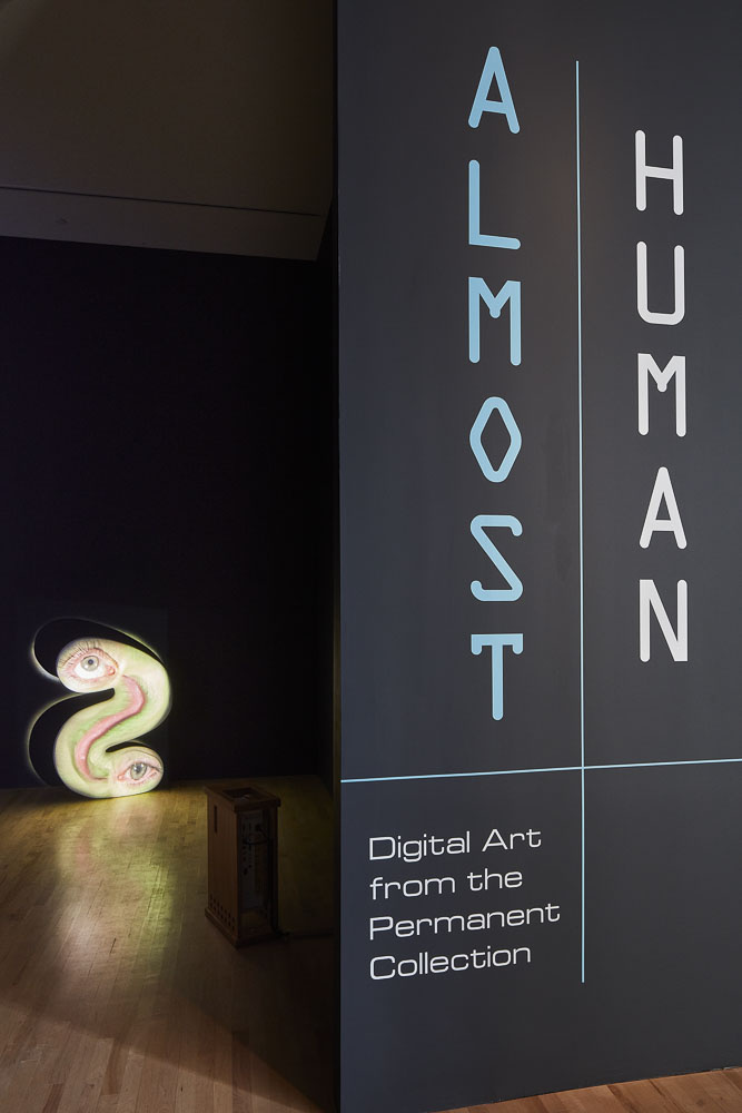 A gallery with black walls and wood flooring. Text on the right reads, “Almost Human: Digital Art from the Permanent Collection.” Inside the gallery is a greenish digital sculptural work shaped like a backwards "S"  with two eyes on either end and an open mouth between them.