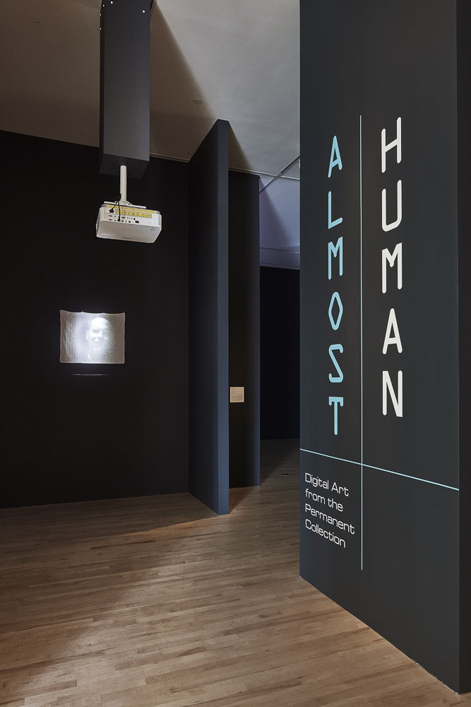A gallery space with black walls and wooden floors with text that reads, “Almost Human: Digital Art from the Permanent Collection.” On the left, a human face seemingly in agony is projected onto a small white screen against the black wall.