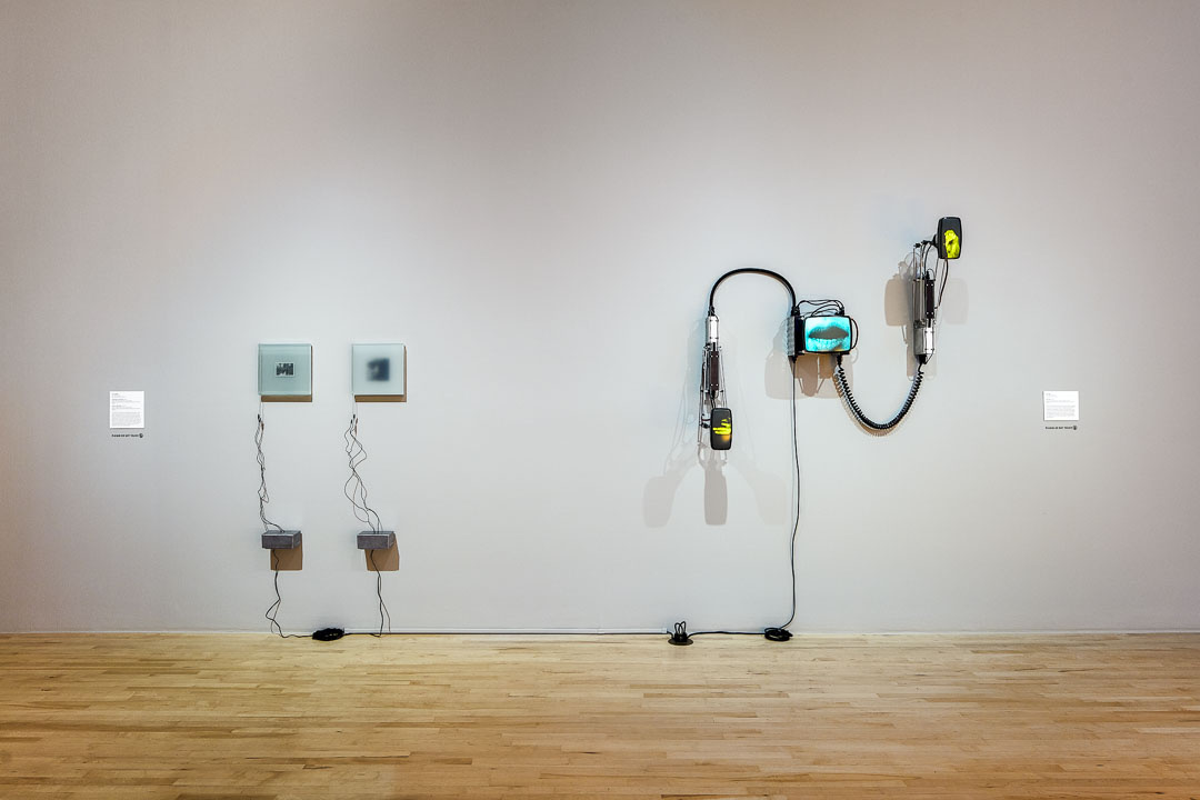 Two digital artworks hang in a gallery. On the left are 2 small square panels with suspended electronic wires. On the right are 3 digitalized screens that are positioned diagonally; linked with attachments. Both works have electric cords plugged into the floor's outlet.