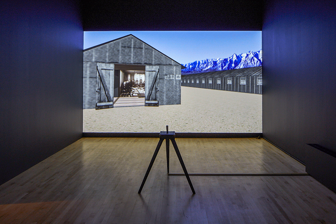 In a dark room a brightly projected image fills the back wall. A black and white barn is erected against sandy ground and a blue sky. Inside the barn is an antique photo of people sitting around a table. In front of the projection is a stool with a video game console.
