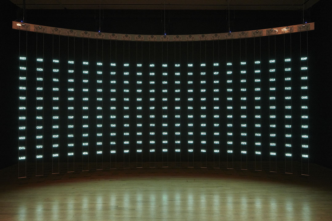 In a dark gallery space, a semi-circular board is suspended with 21 columns that each have 11 rectangles hanging vertically, with words on each rectangle. ​Words are illuminated and the area is spotlighted as the background is completely dark.