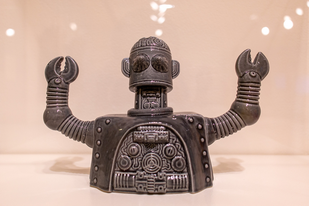 A grey ceramic robot has its arms raised. Its open-ended claw hands and expansion joints are made of ceramic. The robot's raised head is open, revealing a brain. Its interior chest cavity is visible, revealing mechanical looking objects.  
