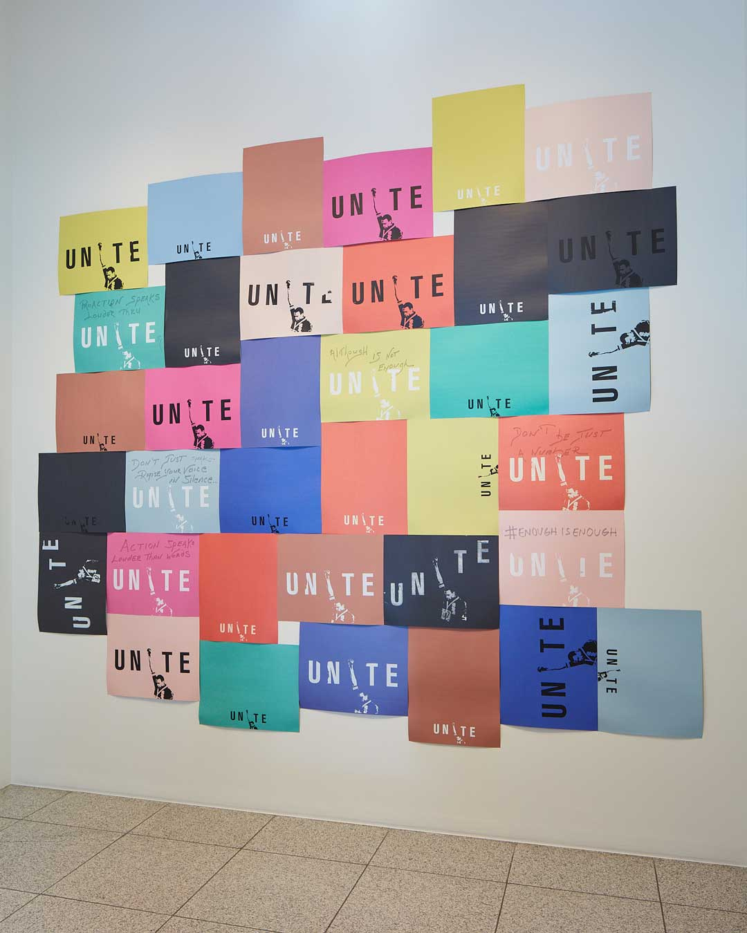 Thirty five pieces of rectangular, multi-colored paper posted on a wall with the word UNITE printed on them. Several have a man with his head lowered while raising his arm and fist high.