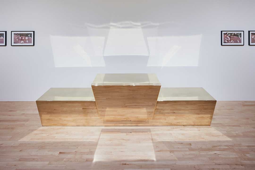 A large, shiny golden colored sculpture, akin to a platform for awards ceremonies with three winners. The golden sculpture reflects the wooden floor of the gallery. Behind the sculpture are four paintings, two on each side.