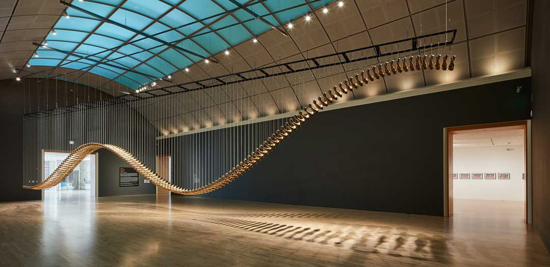 A large gallery with a sculpture hanging low from the ceiling that looks like a long moving snake-like figure.