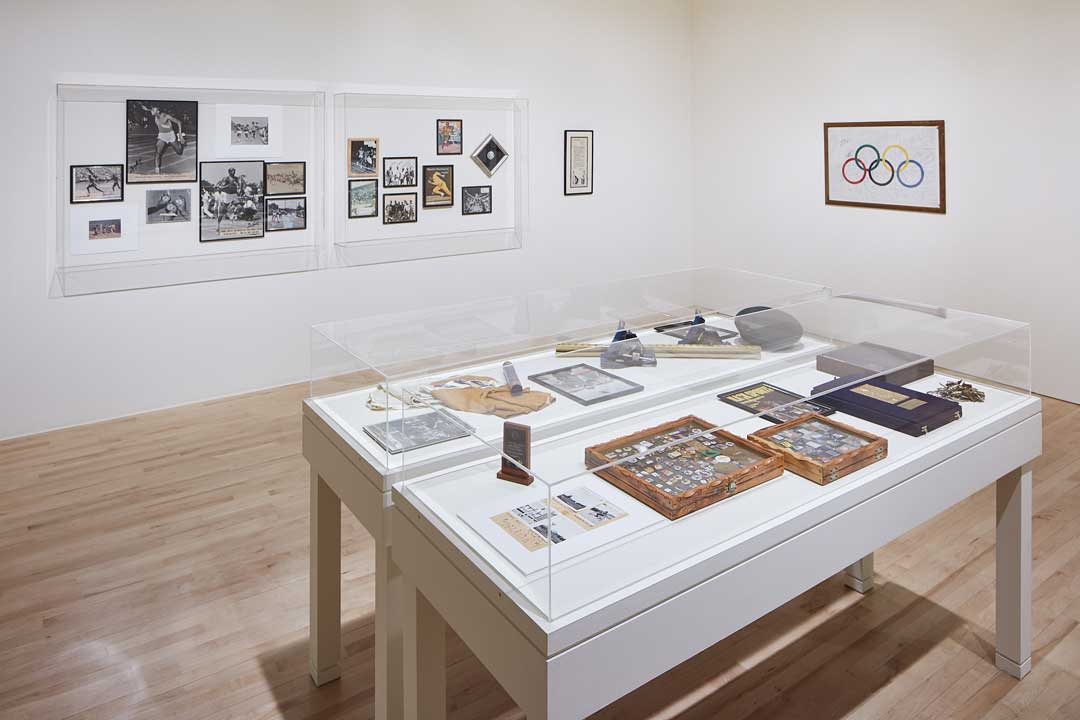 Two display cases with pictures inside on one wall with a separate picture next to it. On another wall is a picture of the Olympic Rings and in the center of the gallery are two side-by-side standing display cases with many different items in each case.