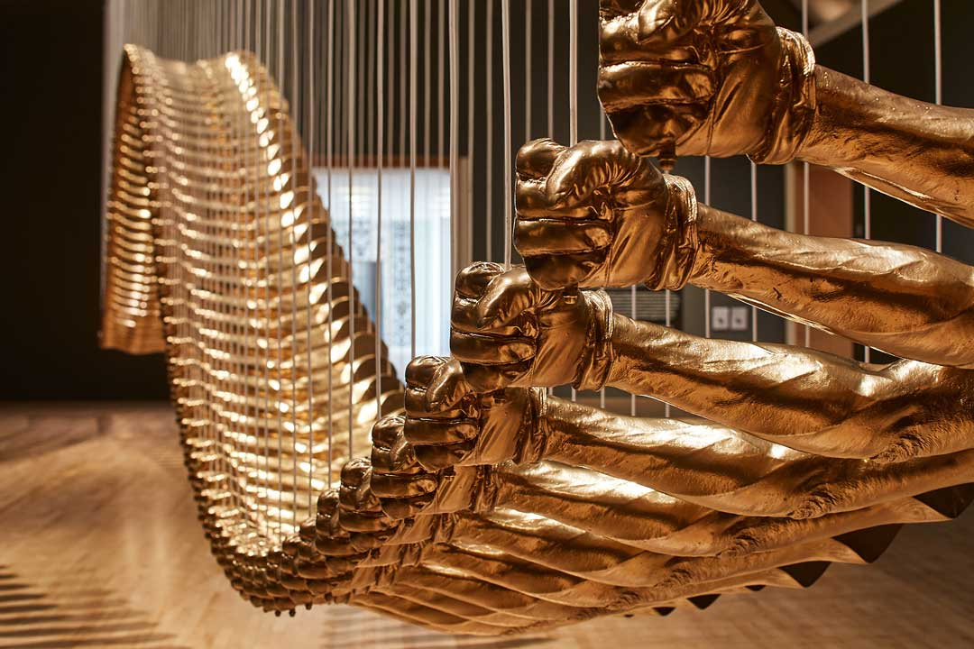 A large wavy sculpture hanging from the ceiling by heavy chords which are holding the beams or slats of the structure. Each beam looks like a strong arm with a closed fist on both sides of the beam.