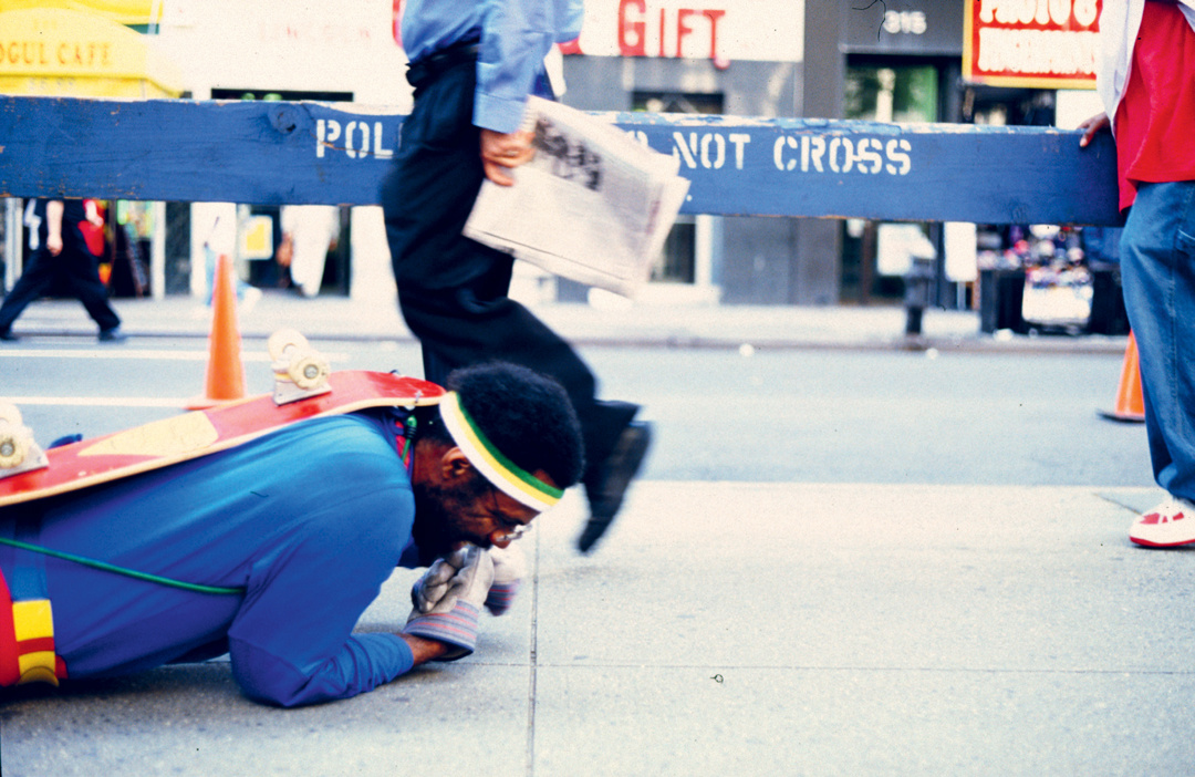 A black man dressed as a super hero lays on the ground with a skateboard strapped to his back. He wears glasses and gloves. He  struggles to move forward. Behind him is a police line barrier against the cityscape. People walk in front of him, visible from the waist down.