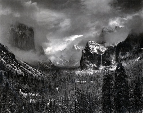 An aerial view of a forest in a distance. Beyond the forest are large mountains, with clouds filling the sky and the space of the photograph. The black and white image looks ominous, with the promise of a storm coming or the aftermath of a fire.