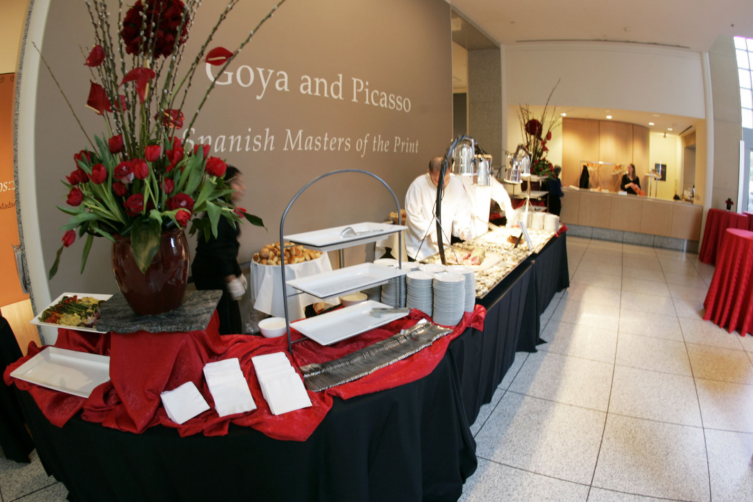 Red tulips sit atop a formal banquet table. Rectangular ceramic plates hold vegetables and 2 catering staff prepare more food at the end of the table. An exhibition title wall with text is behind the table. SJMA’s front desk is in the distance.