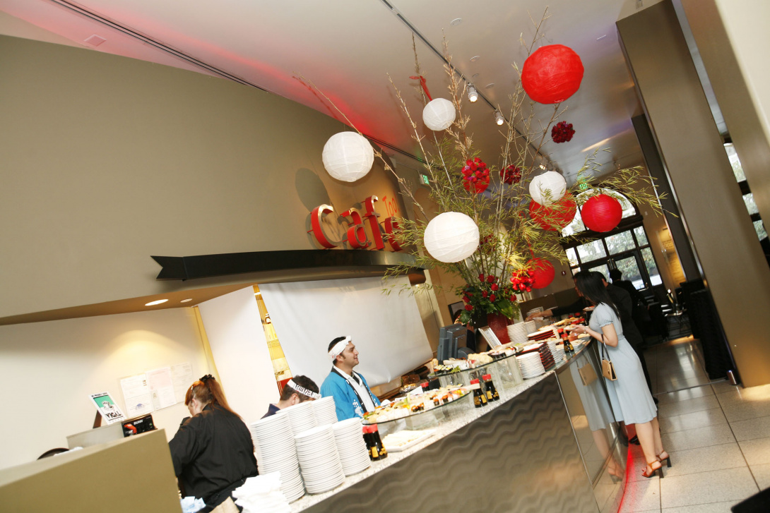 Visitors serve themselves from a buffet in a café. A large bamboo plant and red and white lanterns decorate the space. 3 catering staff are behind the buffet. A large overhead sign reads “Café” and a glass door to the street is in the distance. 