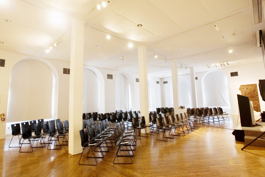 Roughly sixty black chairs face a podium at the front of a room with white walls, white columns, and wooden floors.. A black podium with a microphone is at the front of the room.  A red fire extinguisher is on the wall to the left of the chairs. 