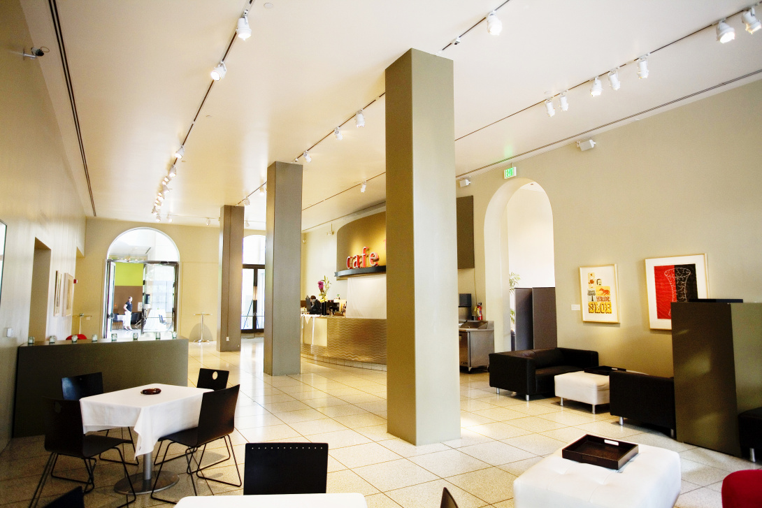 A look into SJMA’s café, facing towards the main museum entrance. Small square tables with tablecloths and chairs each punctuate the space. Three columns are in the middle of the room. The word “cafe” is in three-dimensional letters above a counter. 
