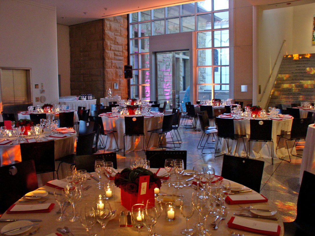 A lobby is set with elegant tables featuring white tablecloths, red accent napkins, wineglasses, candles, and roses. There's a staircase in the background and an elevator to the left. A glass window behind the tables reveals an outside view to the street.