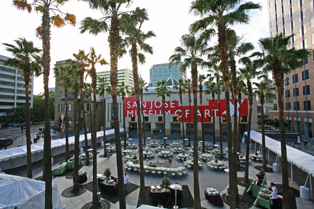 Outdoors, the San José Museum of Art is visible through a center of palm trees. There are large white tents in surrounding round tables with green table cloths and chairs. There are servers standing behind a small bar.