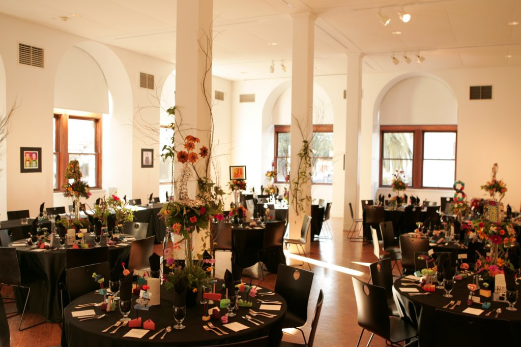 Inside a white room with red and orange flowers with vines are placed on the walls and pillars of the room. There are round black tables with flowers, place settings, silverware, and glassware on the tables. Arched windows are open with sunlight shining through.