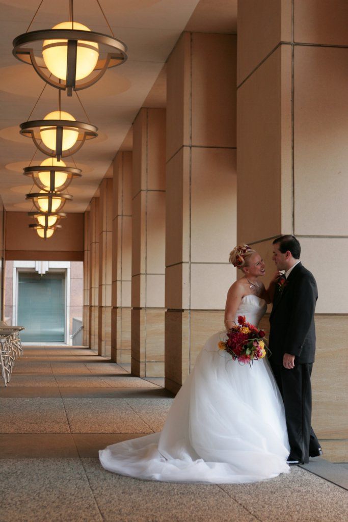 A man wearing a black suit stands next to a woman wearing a white dress holding a bouquet of flowers. They are smiling at each other in a hallway outside the museum.