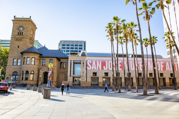 The exterior of a long building that has a large red banner that reads “San Jose Museum of Art.” A circle of palm trees are to the left of the building, with the historic clocktower visible to the far end of the museum.