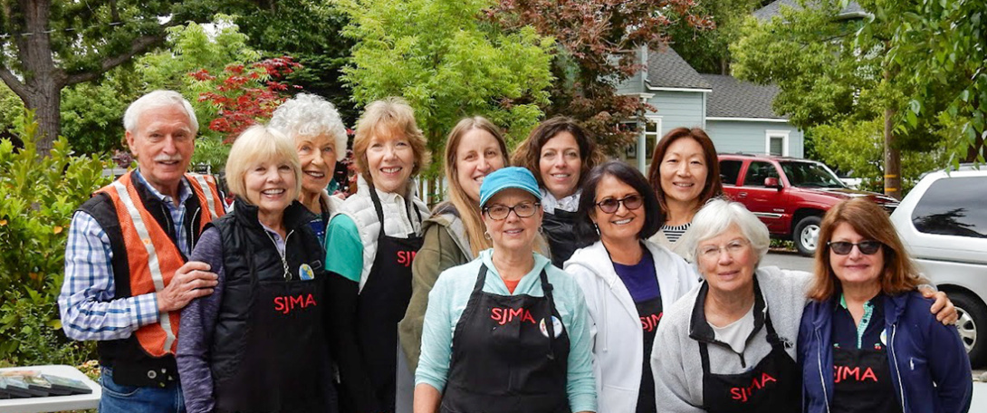 A group of smiling SJMA Museum Store Guild members posing together outdoors, wearing aprons with the SJMA logo.