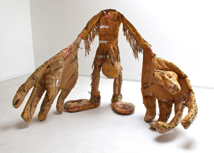 A cardboard sculpture of a man with no head. The hands and feet are unusually large and disproportionate to its body. The arms, legs, and torso are covered with tassels (like a cowboy's). In between his legs is a round object, almost like a small pouch.