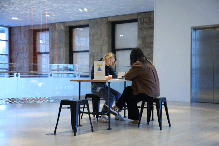 Two people write at a table in a gallery, with a sculpture hanging on a stone wall behind them.